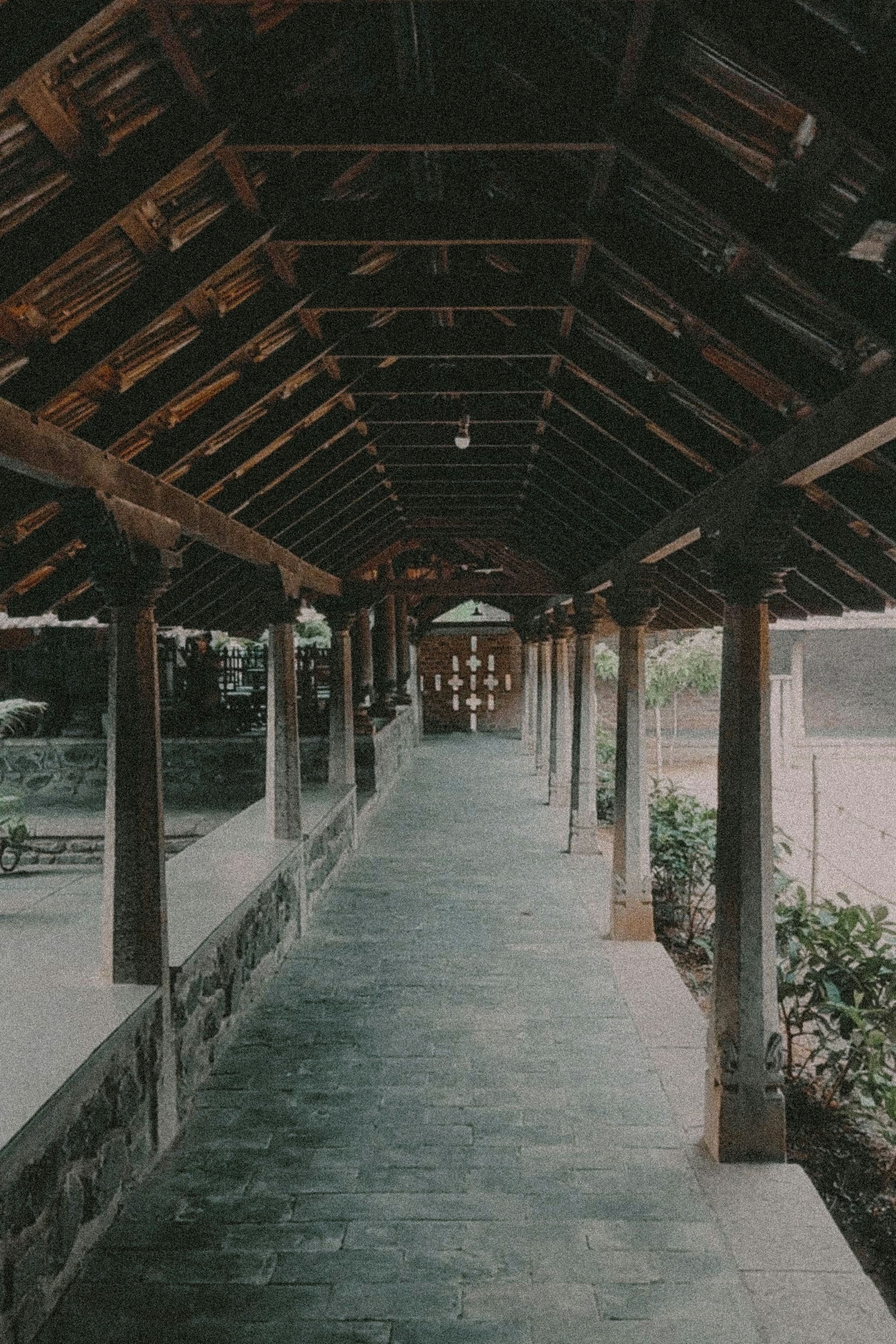 A covered walkway in traditional Kerala style, timber rafters over a stone floor and low laterite wall.